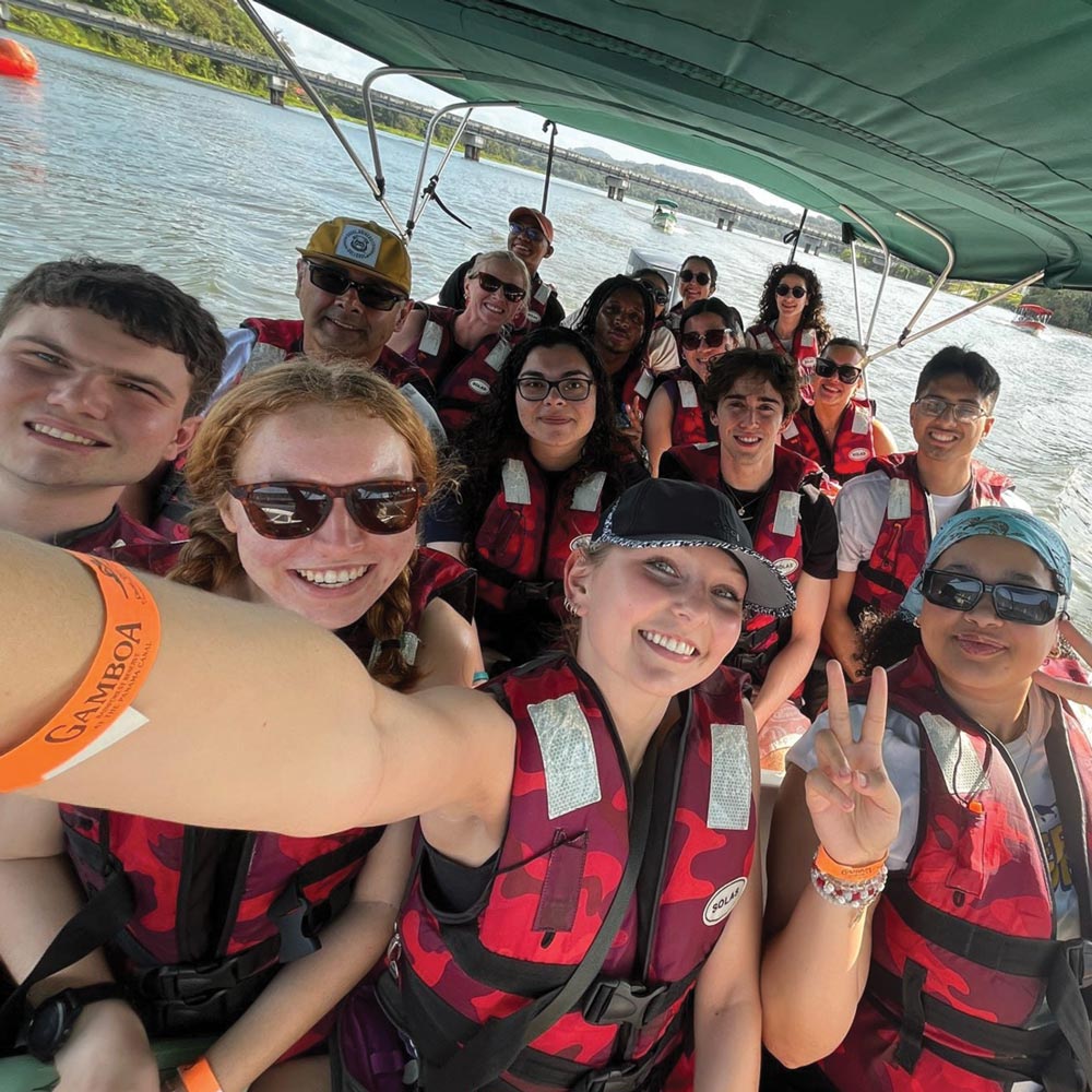 A group of people in life jackets on a boat under a green canopy, smiling and posing for a selfie.