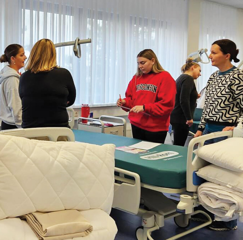 Five people in a hospital room with hospital beds, one writing on a clipboard.