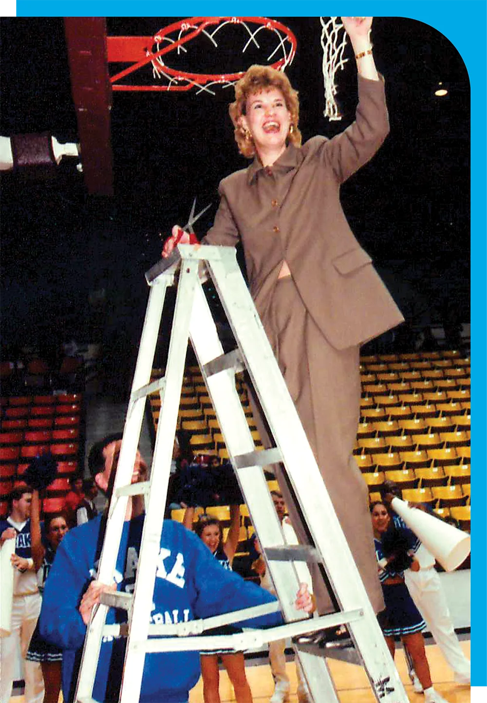 Woman on a ladder cutting down a basketball net, with cheerleaders and a stadium in the background.