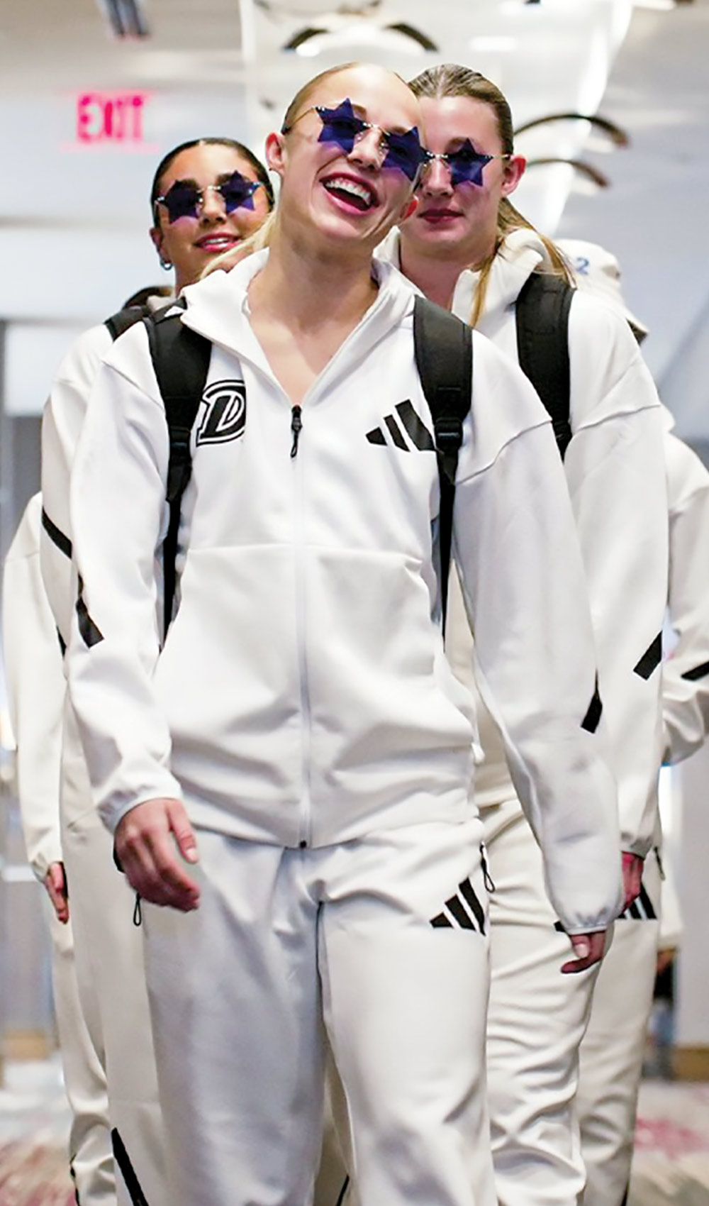 A group of individuals in white tracksuits and star-shaped sunglasses walking in a corridor.