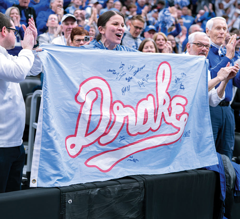 Fans cheering with a signed "Drake" flag at a sporting event.