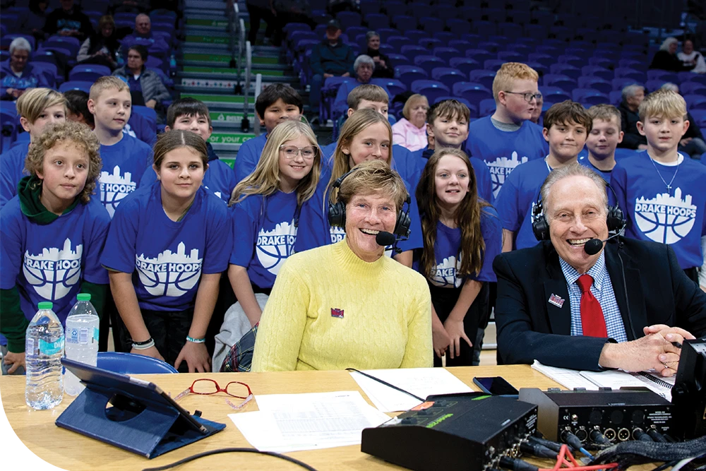 A group of children in blue "DRAKE HOOPS" T-shirts stands behind two adults with headsets at a commentary table.