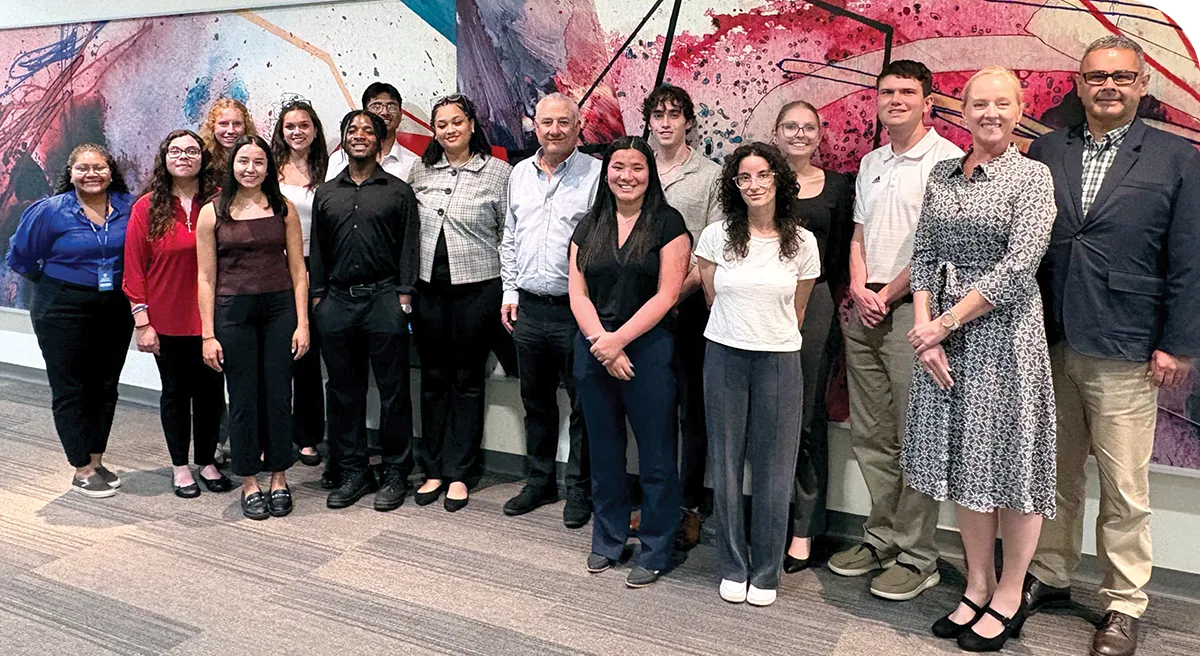 A group of fifteen people stands in front of a colorful abstract mural, wearing business casual attire.