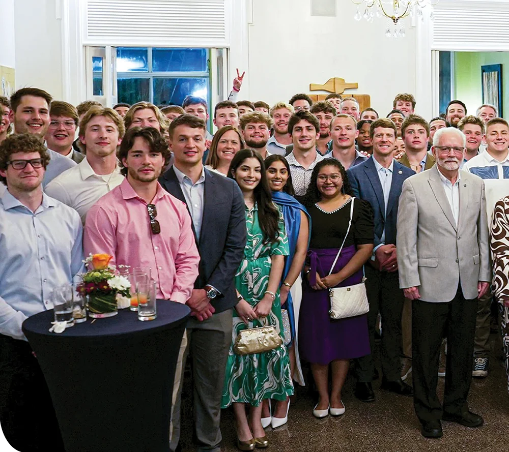 A large group of people in semi-formal attire at a social event indoors with windows, a cross, and a cocktail table with drinks.