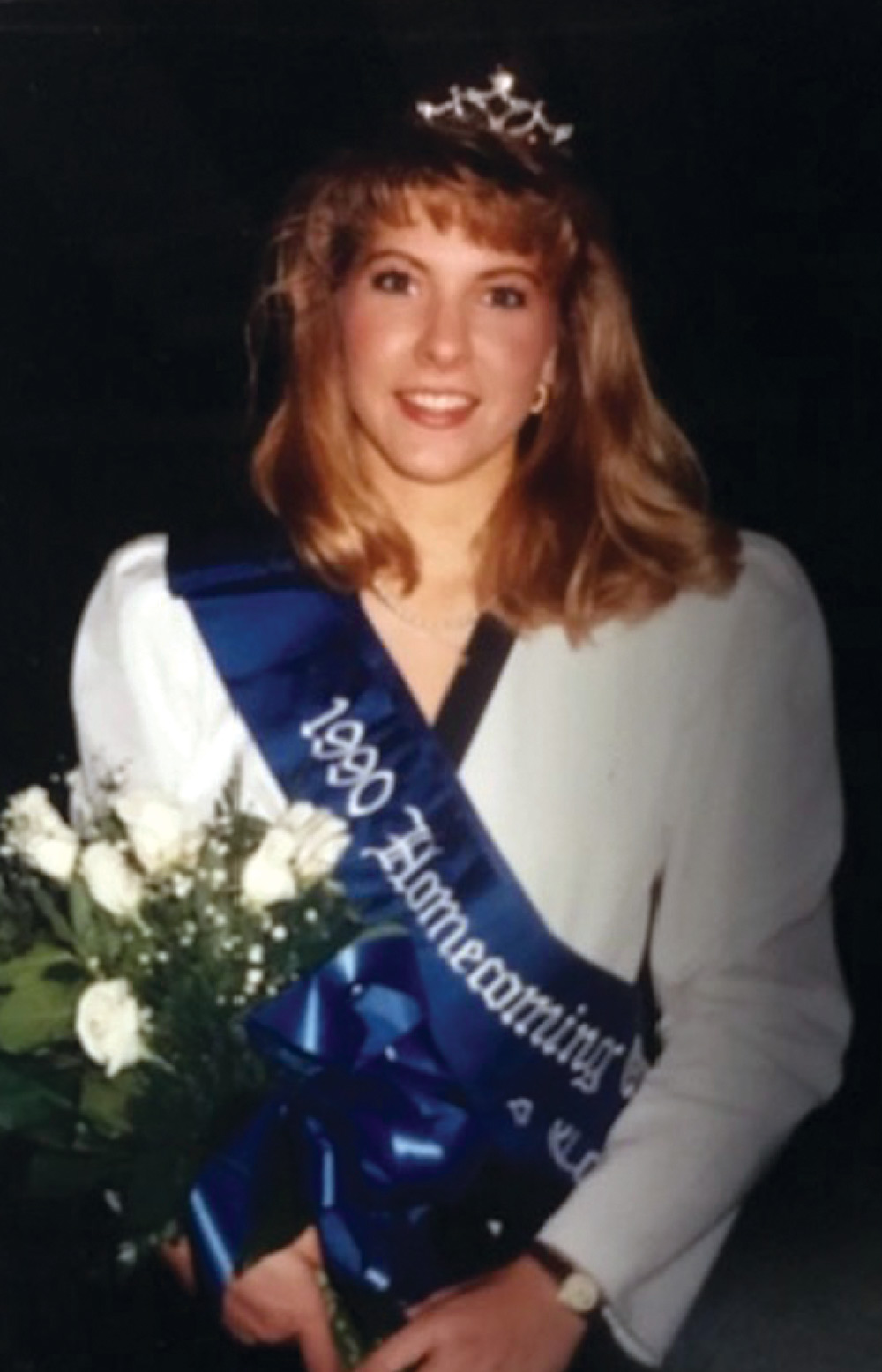 A person wearing a tiara and a "1990 Homecoming" sash, holding white roses.