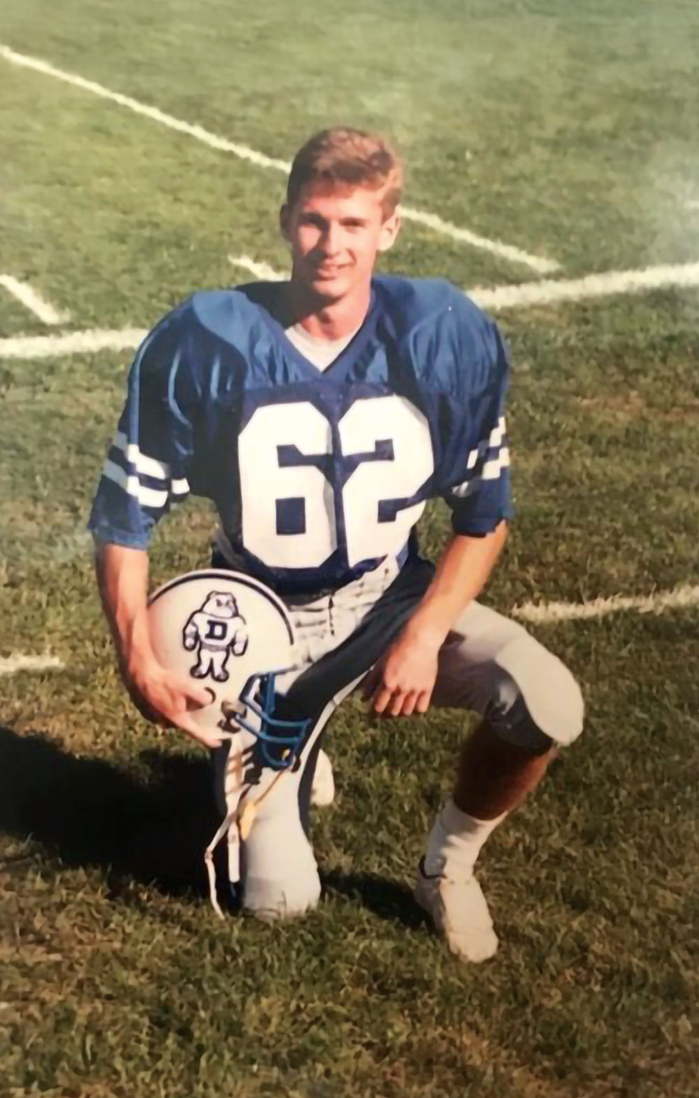 A young man in a blue football jersey with number 62, kneeling on a grassy field, holding a helmet with a bulldog and the letter D.