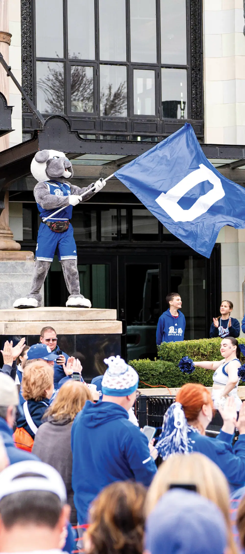 Mascot waving a blue flag with the letter "D" in front of a cheering crowd.