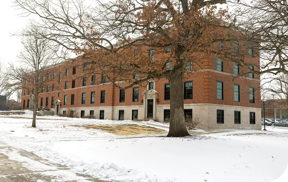 A large, three-story brick building with many windows, surrounded by snow and leafless trees.