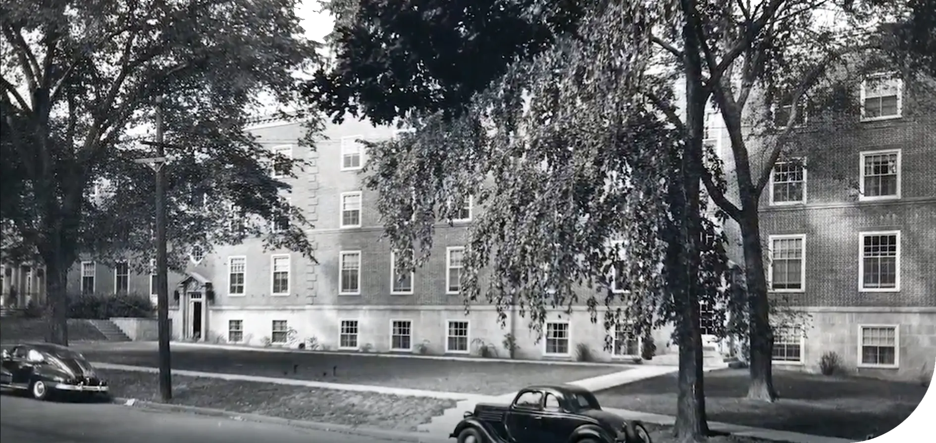 Black and white photo of a four-story brick building with vintage cars and trees in the foreground.