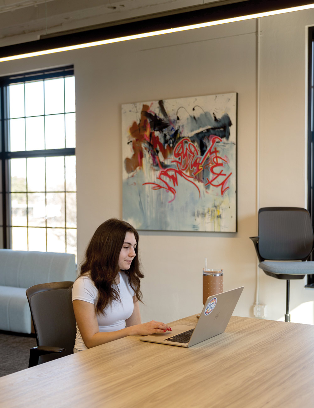 A woman working on a laptop at a wooden table with abstract art in the background.