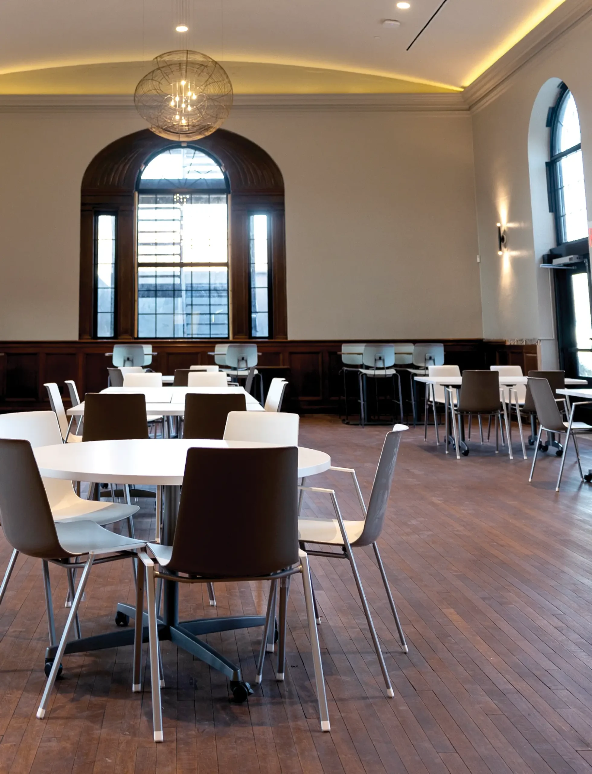 A spacious room with round white tables and a mix of white and dark brown chairs, featuring large windows and a spherical light fixture.