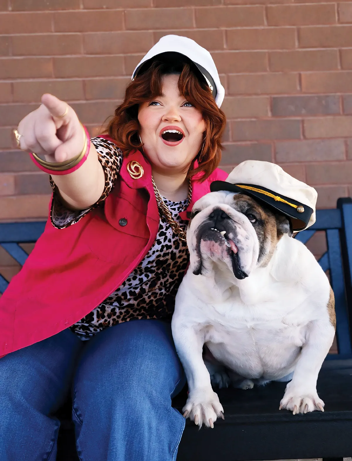 A person pointing excitedly while sitting next to a bulldog wearing a sailor's hat on a bench.