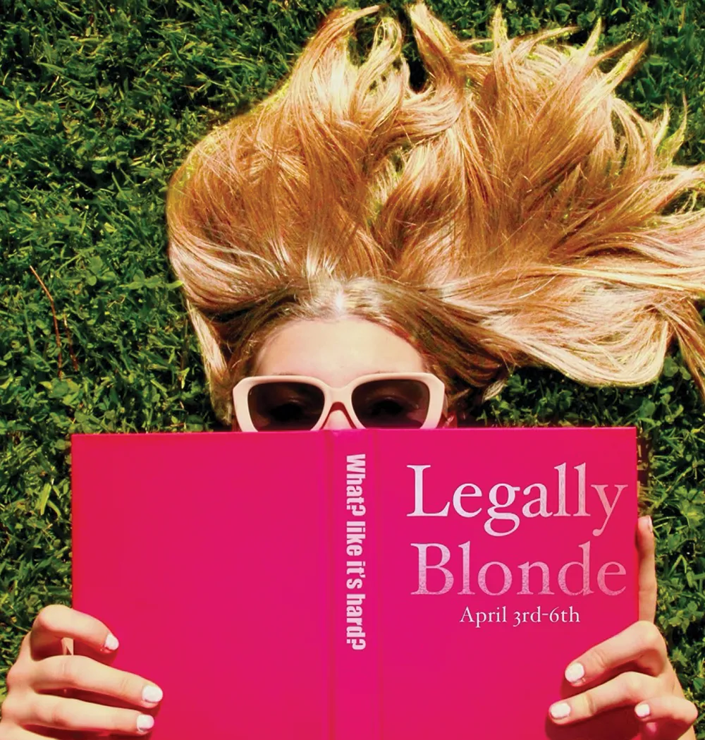 Person with long blonde hair and sunglasses reading a pink book on grass.