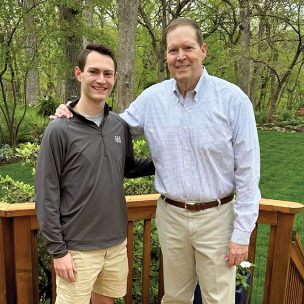 Two men standing together on a wooden deck in front of a lush garden.
