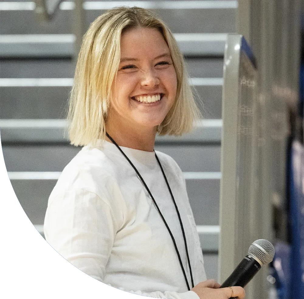 Person with short blond hair smiling and holding a microphone indoors.