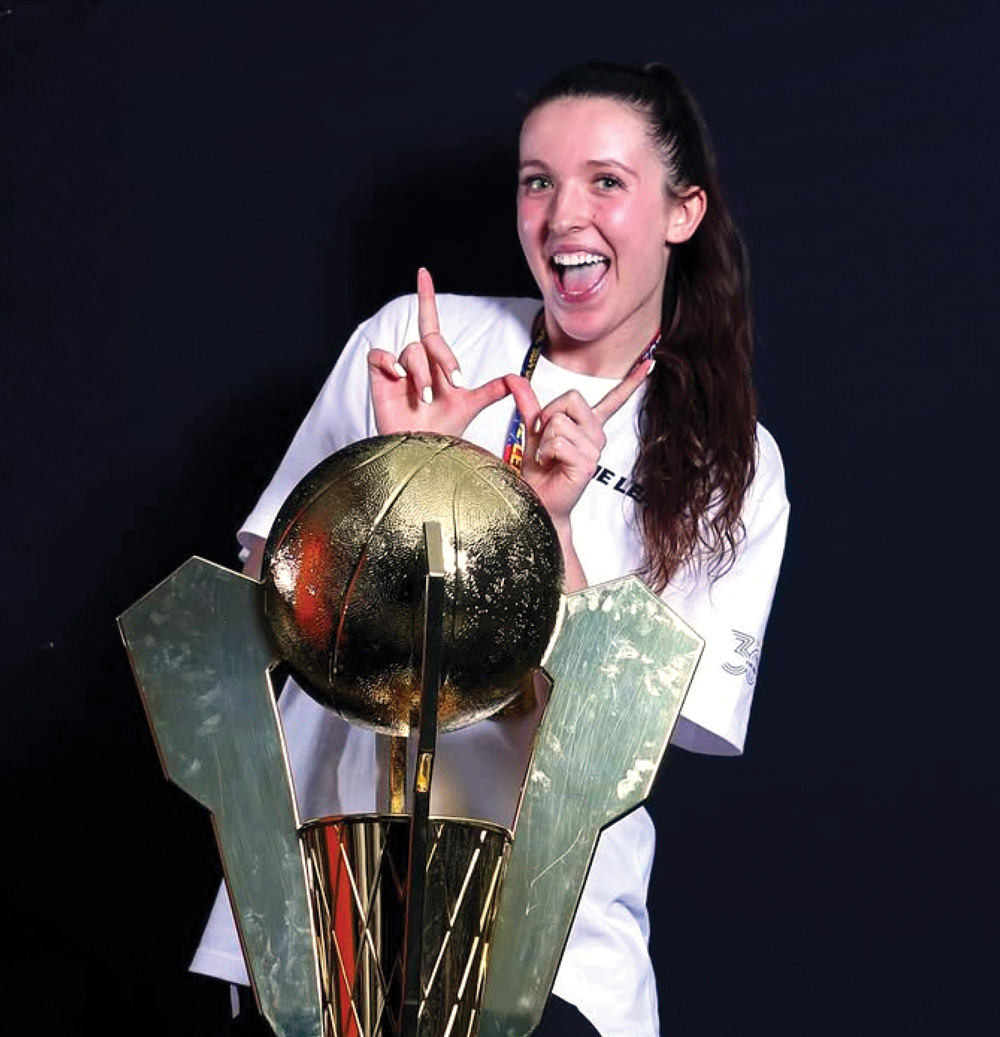 Person smiling behind a large gold trophy with a black background.