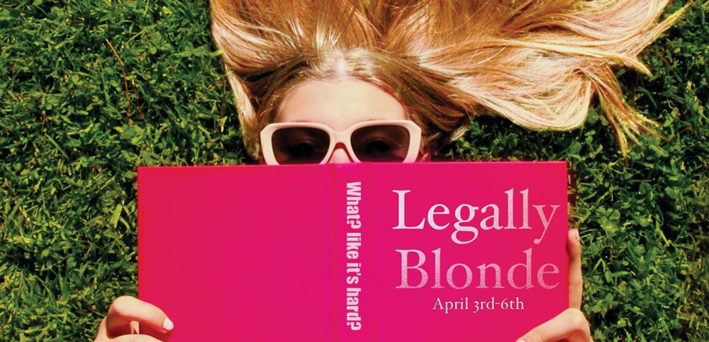 Person lying on grass, holding a pink book titled "Legally Blonde," with sunglasses and hair spread around.