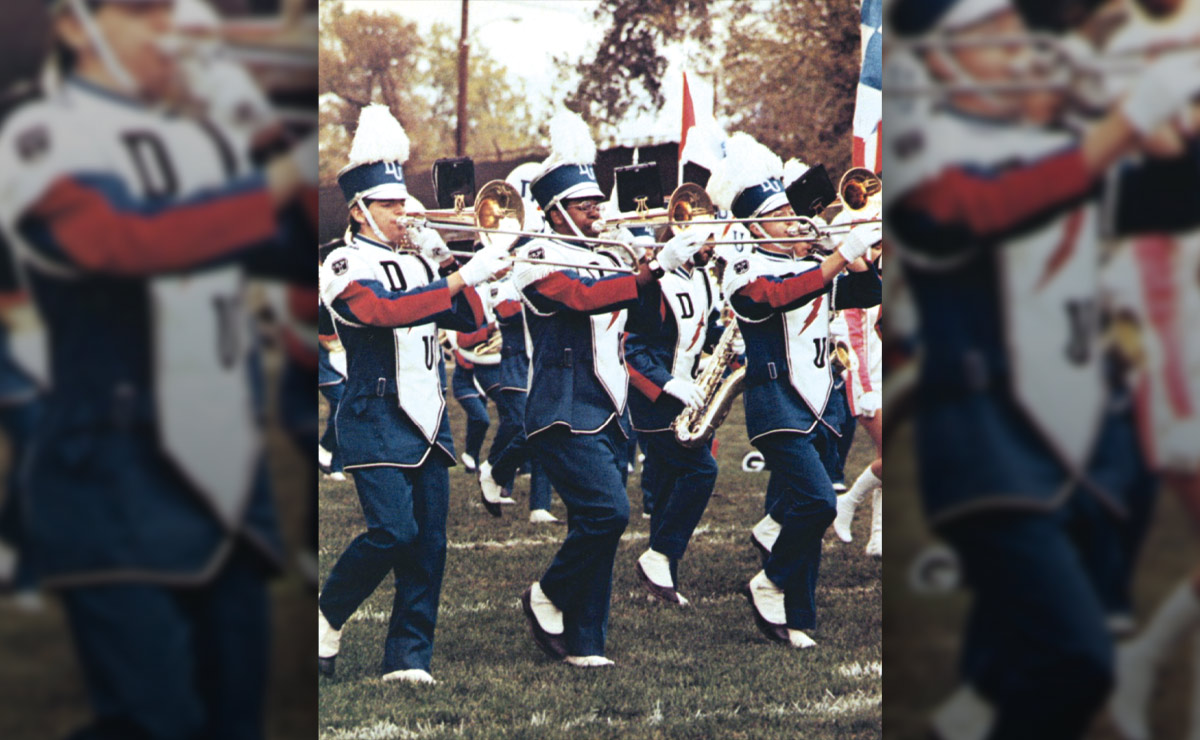 Marching band members playing trombones, wearing blue and white uniforms with plumed hats.