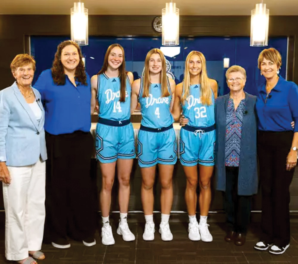 A group of seven women, including athletes in light blue basketball uniforms and individuals in formal attire, stand in a row.