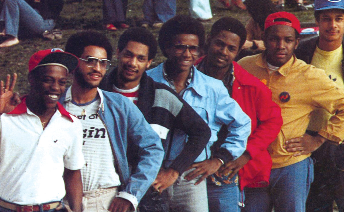 A group of seven men posing diagonally, all wearing casual jackets and caps in various colors.