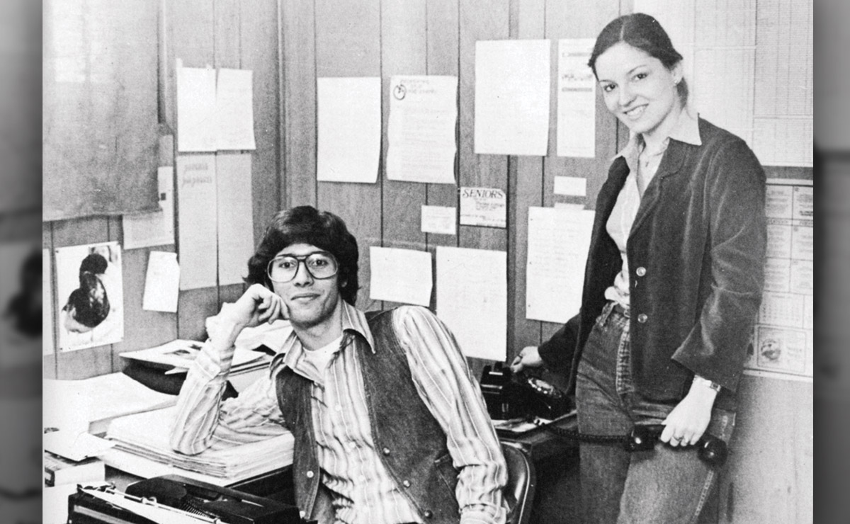 Two people in an office with papers and documents on the wall behind them.