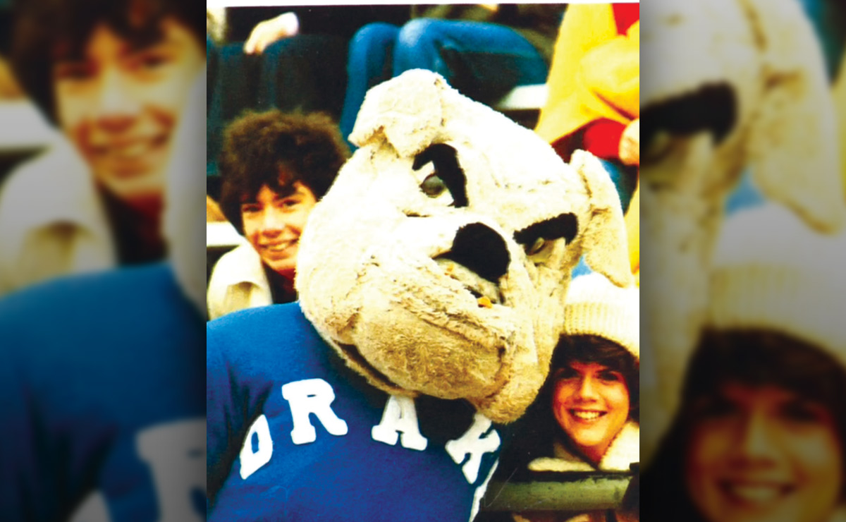 A person wearing a bulldog mascot head with "DRAKE" on their blue shirt, surrounded by two people in a stadium setting.