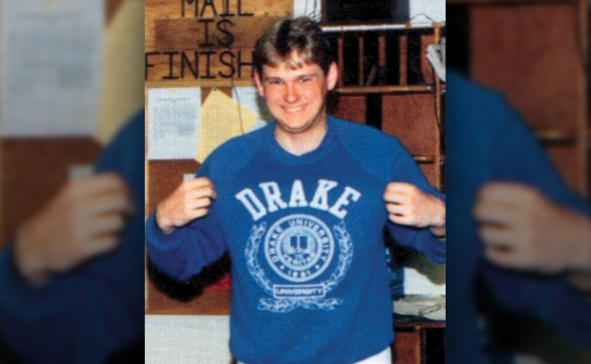 Person wearing a blue "DRAKE UNIVERSITY" sweatshirt, smiling with a background of wooden shelves and a sign that reads "MAIL IS FINISH".