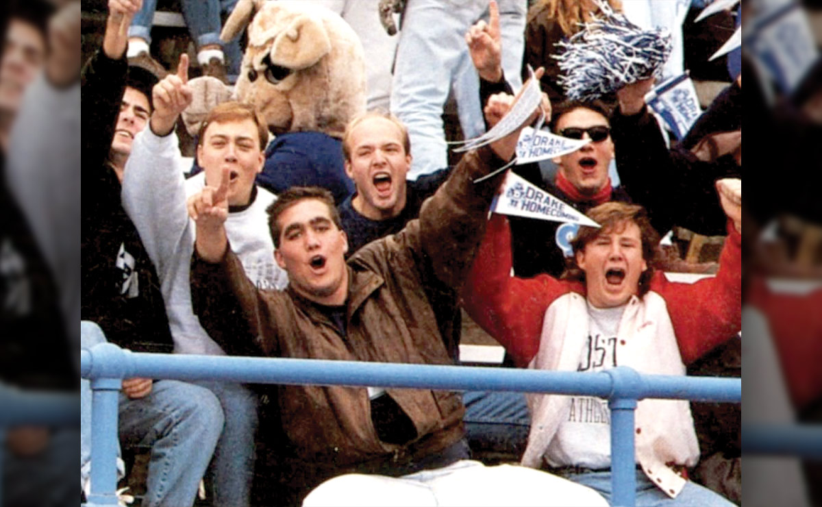 Group of enthusiastic fans cheering at a sporting event with some holding "Drake Homecoming" pennants.