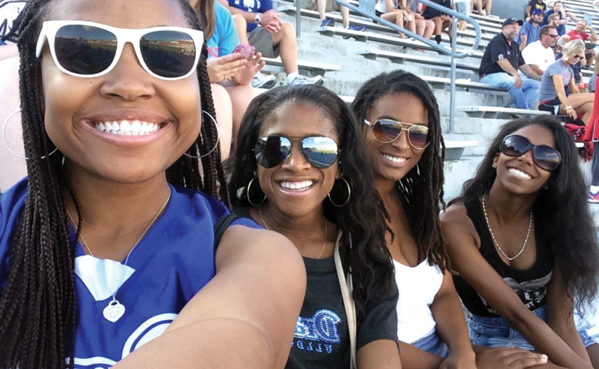 Four young women smiling in a stadium, wearing sunglasses and casual attire, with spectators visible in the background.