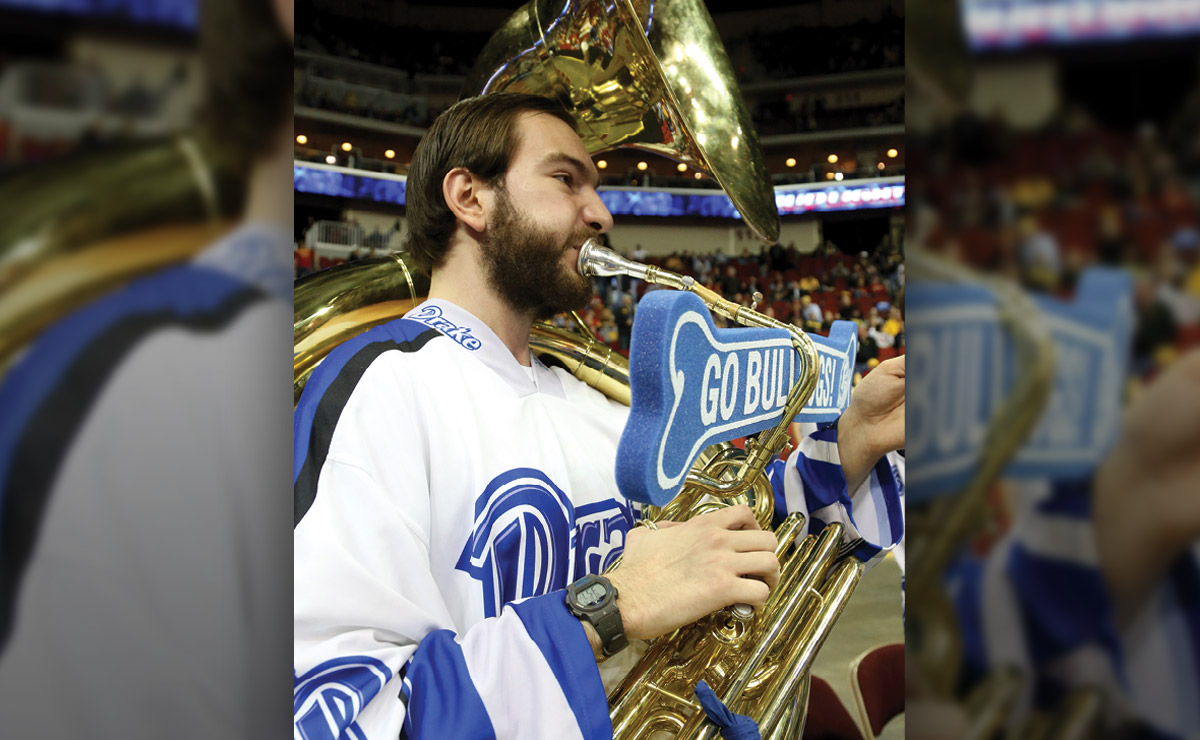 Musician playing a sousaphone in a sports arena with a foam sign attached.