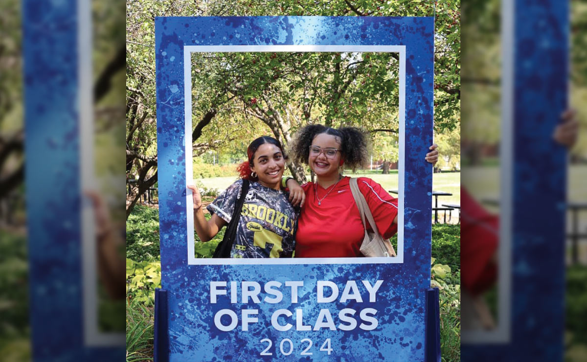 Two people smiling, framed by a large blue display with trees in the background.