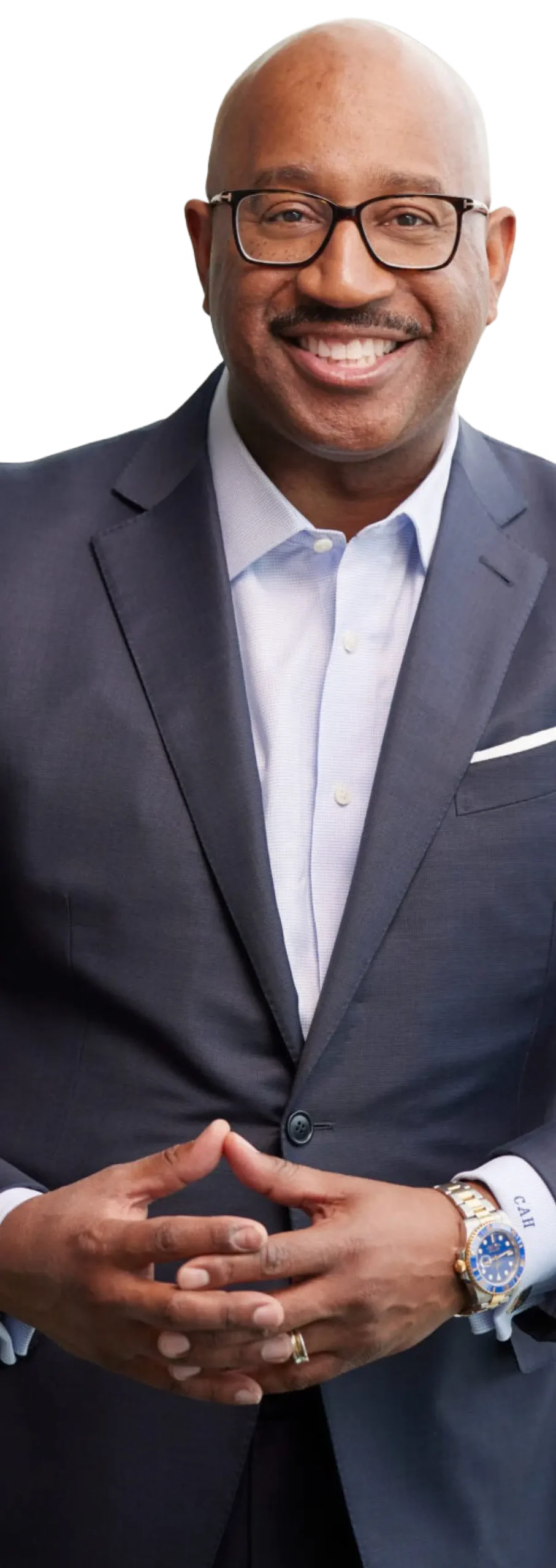 Cain A. Hayes, a smiling bald man with prescription eyeglasses, a dark navy blue blazer business suit, light sky blue button-up dress shirt underneath, and a silver watch on his wrist as his hands are interlocked together