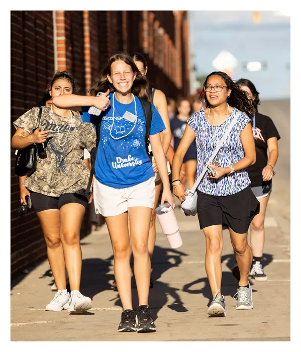 Group of incoming students walk down a sidewalk near campus buildings, smiling and talking, wearing casual clothing and Drake University apparel during move-in or welcome activities.