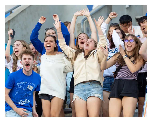 Group of Drake University students cheering with raised arms in stadium seating, smiling and celebrating during a campus event or athletic game.