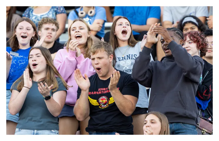 Crowd of students clapping and cheering from bleachers, wearing casual clothing and Drake University shirts, reacting enthusiastically during a campus or athletic event.
