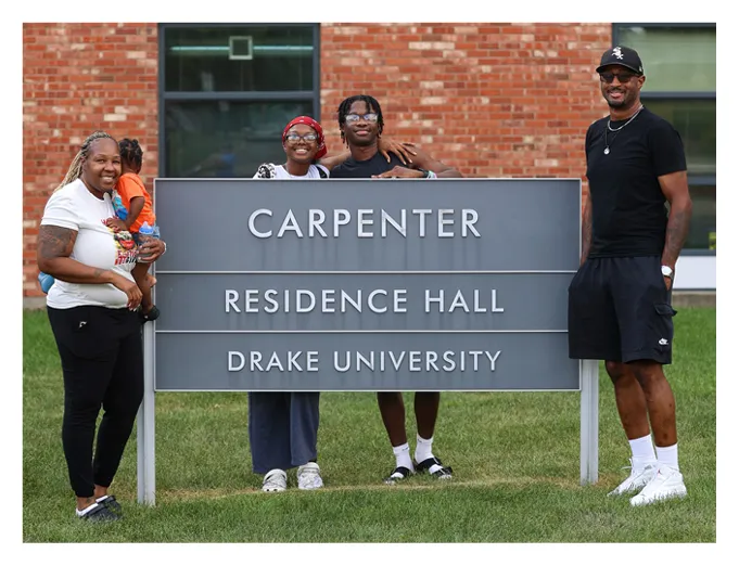 Four people pose outdoors beside a sign that reads “CARPENTER RESIDENCE HALL, DRAKE UNIVERSITY,” standing on grass in front of a brick building, smiling for the camera.
