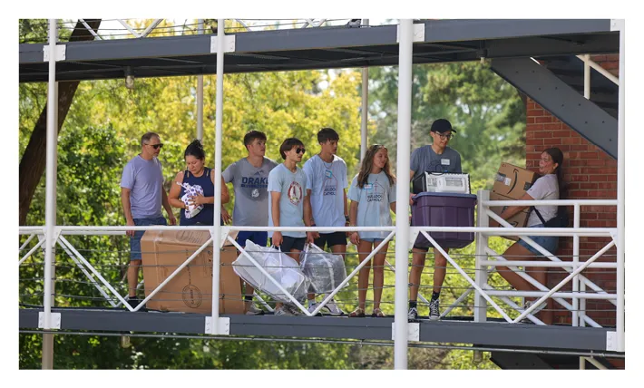 Group of students walk along an elevated outdoor walkway carrying cardboard boxes, plastic bins, and personal belongings during residence hall move-in.