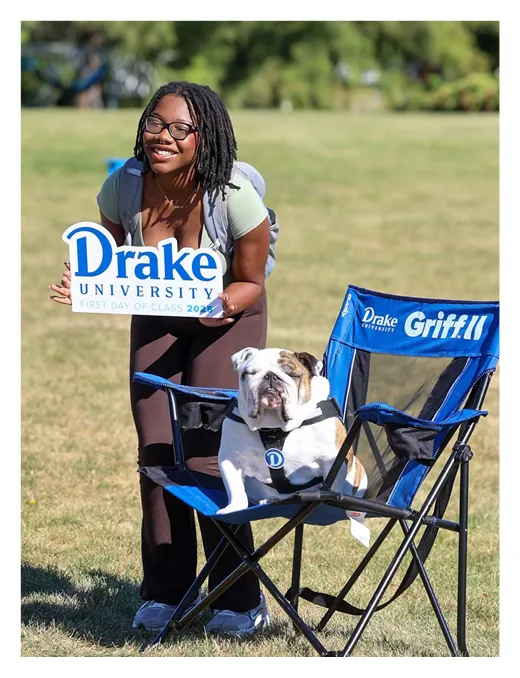 Student smiles while holding a sign that reads “Drake University First Day of Class 2024,” standing on a grassy lawn next to a bulldog sitting in a blue chair labeled “Drake Griff II.”
