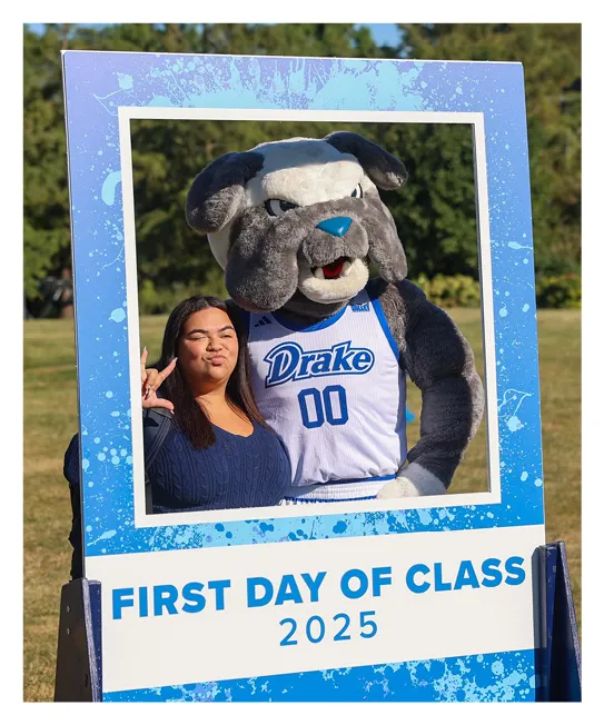 Student poses with the Drake University bulldog mascot inside a large photo-frame prop on a grassy lawn. The frame displays the text “FIRST DAY OF CLASS 2025,” and the mascot wears a white Drake jersey with the number 00 as both smile for the camera.