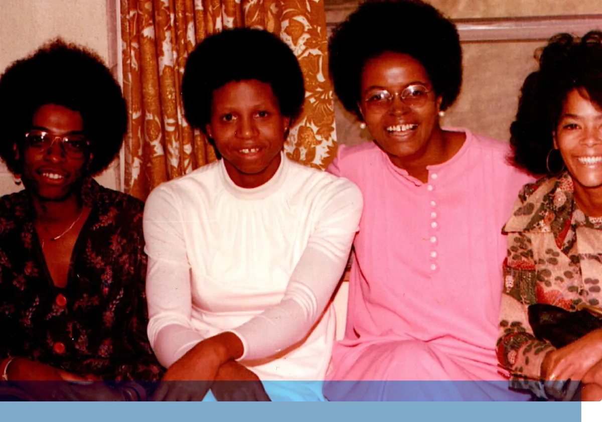 Vintage photograph of four smiling young Black women sitting together in a room with patterned curtains, celebrating friendship and sisterhood.