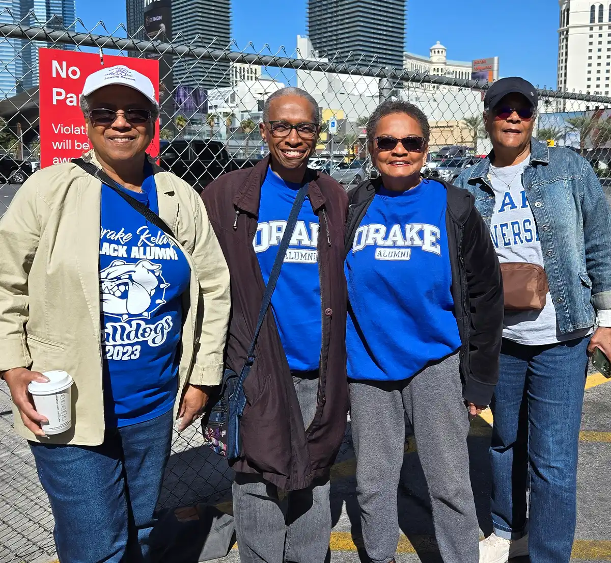 Four smiling Black women wearing Drake University alumni sweatshirts and t-shirts standing together outdoors near a chain-link fence.