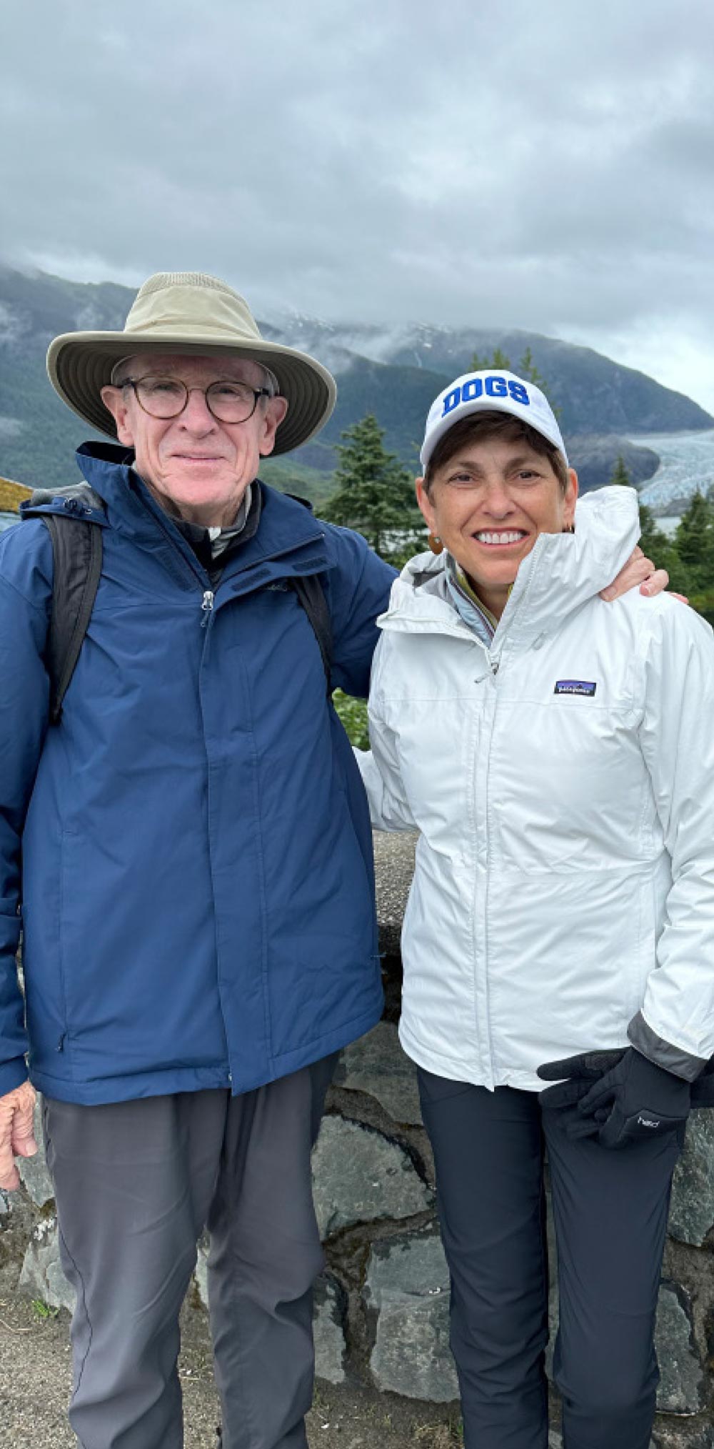 A man in a sun hat and a woman in a "DOGS" cap and white jacket pose together outdoors with a glacier in the background.