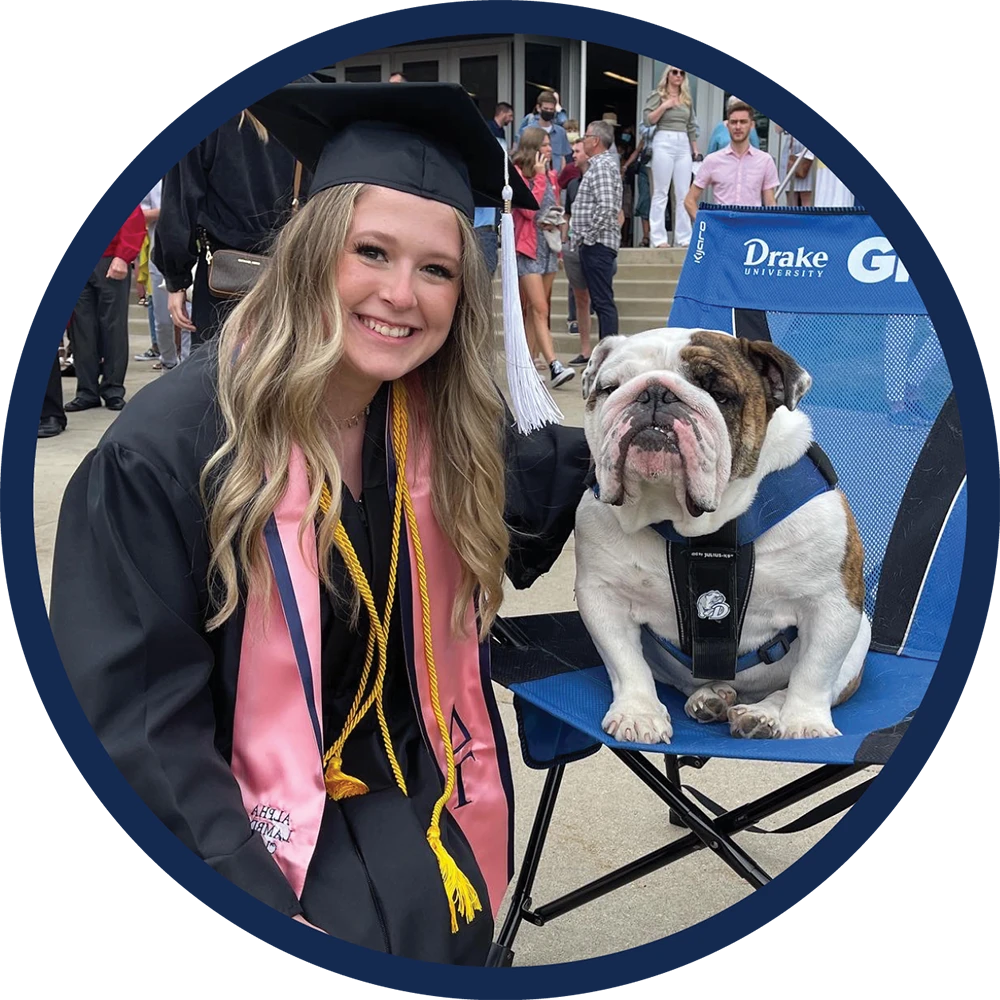 Kayla Hruska in a black cap and gown with a pink stole sits outdoors next to a brown and white English Bulldog resting on a blue Drake University folding chair.