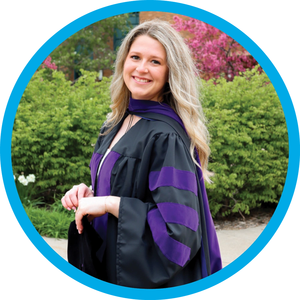 Kayla Hruska smiles while wearing a black and purple doctoral graduation gown, standing outdoors in front of green shrubs and pink spring blossoms.