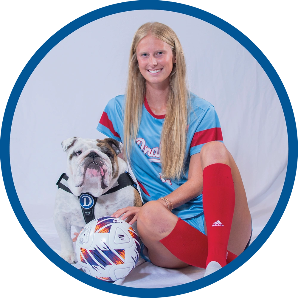 Avery Mertz in a blue and red uniform sits on a white backdrop with a soccer ball and an English Bulldog wearing a Drake University harness.