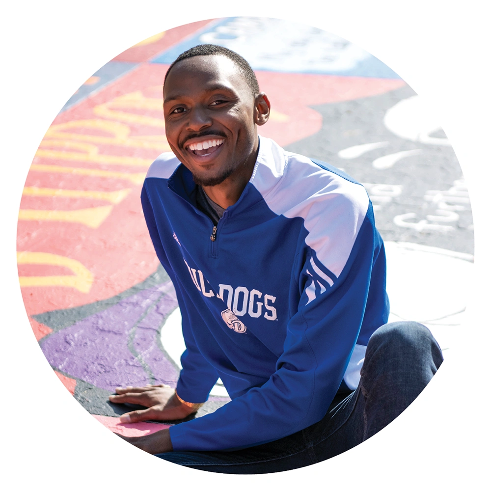 Donovan McCamey in a blue "Bulldogs" quarter-zip pullover sits and laughs on a vibrant, colorfully painted pavement street on campus.