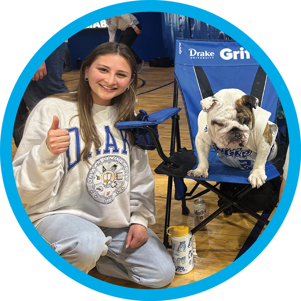 Catalina Samaniego in a white Drake University sweatshirt gives a thumbs up while kneeling next to an English Bulldog sitting in a blue folding chair.