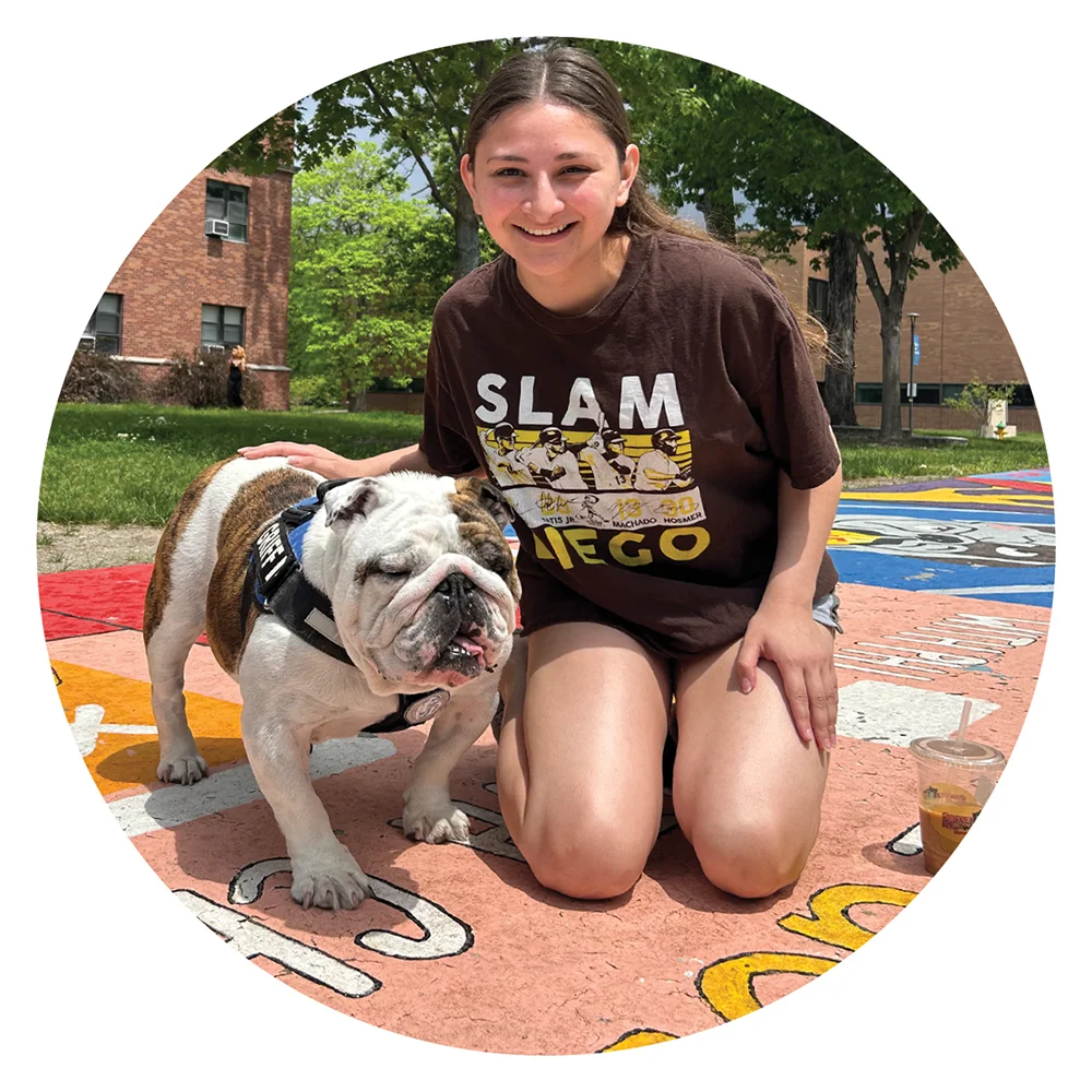 Catalina Samaniego in a brown "Slam Diego" t-shirt kneels on a colorfully painted campus sidewalk, smiling while petting a brown and white English Bulldog.