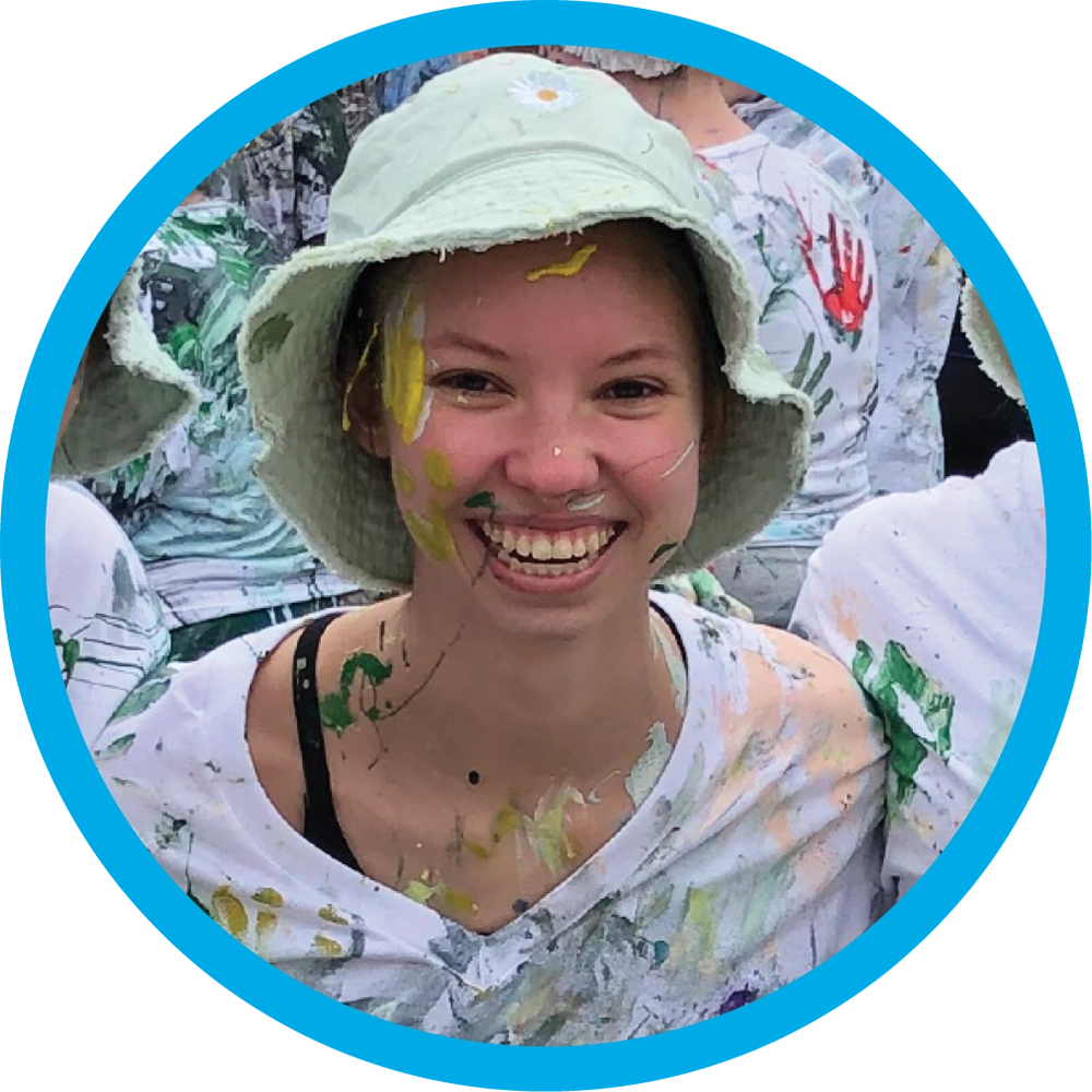 Bridgid Miller wears a light green bucket hat and a white t-shirt, with her face and clothing playfully covered in colorful splattered paint.