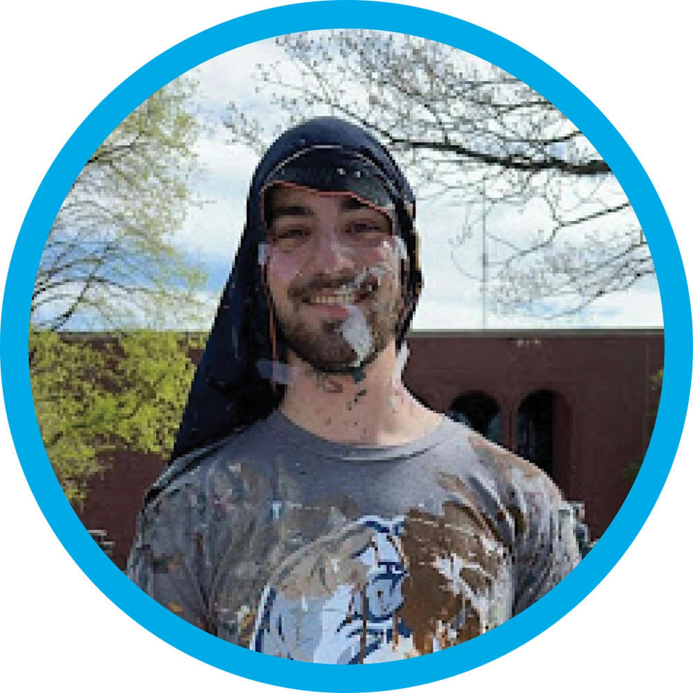 Trevor Stevens stands outdoors with white paint splattered on his face and t-shirt, wearing a dark cloth draped over his head.